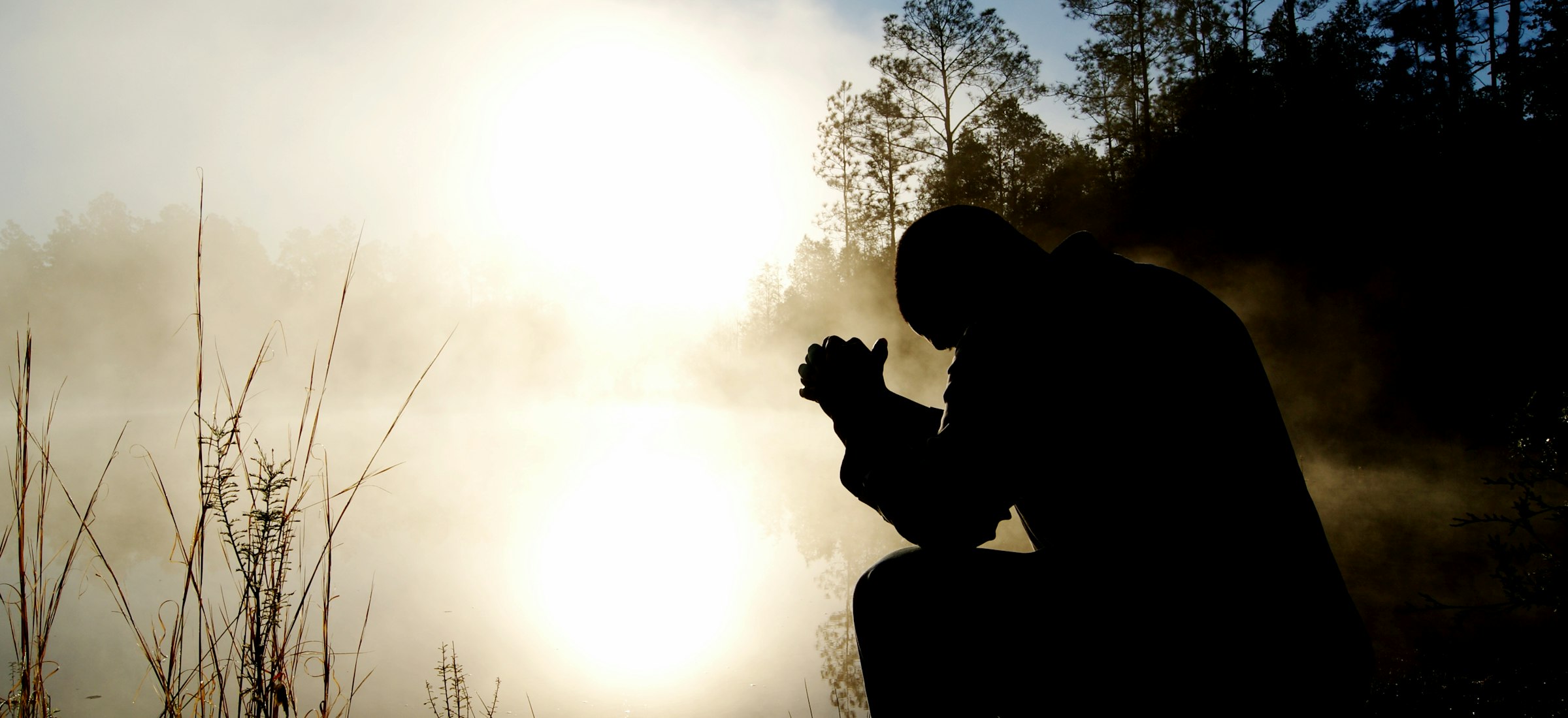 A person kneeling in silhouette beside still water, in mist, against a low sun.